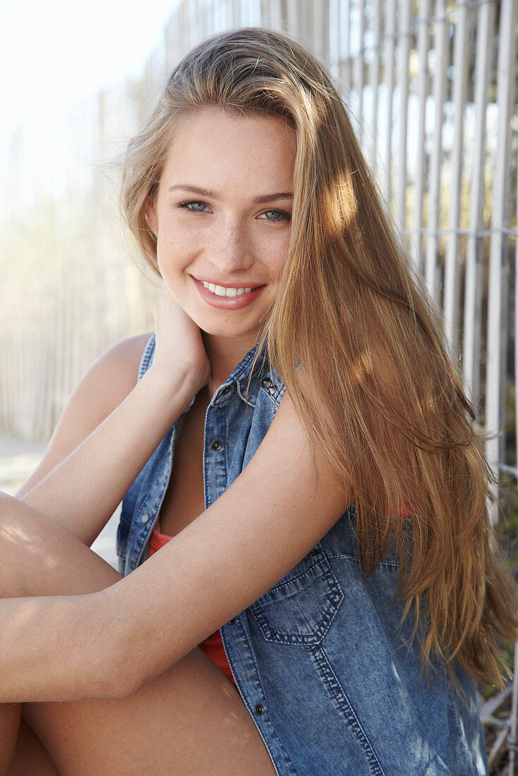 A young blonde woman sitting against a fence wearing a denim jacket