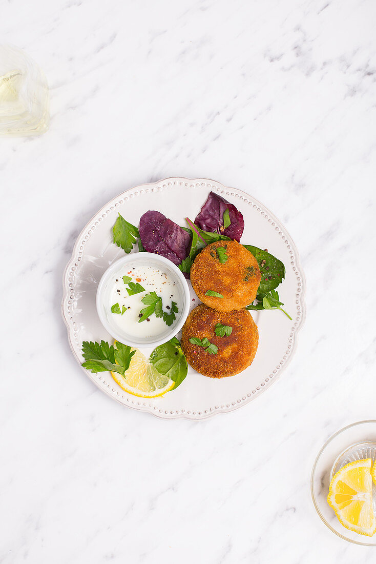 Fish patties on a white plate served with greek yogurt, lemon and fresh herbs