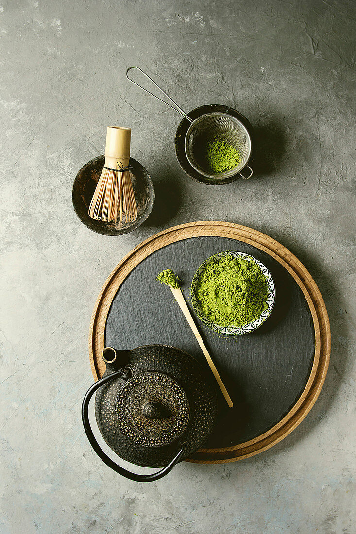 Ingredients for making matcha drink. Green tea matcha powder in ceramic bowl, traditional bamboo spoon and whisk on slate board, black iron teapot over grey texture background