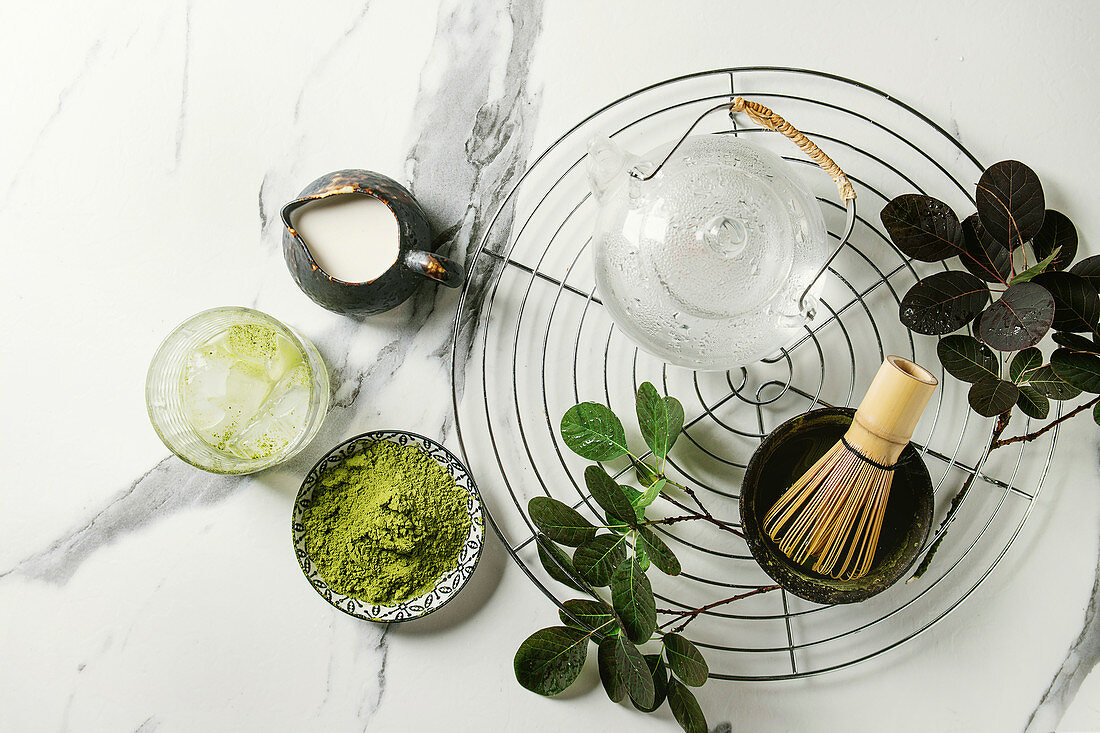 Ingredients for making matcha ice drink. Green tea matcha powder in ceramic bowl, traditional bamboo spoon, whisk on cooling rack, glass teapot, ice cubes