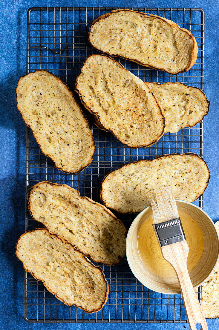 Chargrilled oblong bruschetta bases on a wire rack with a bowl of oil and a pastry brush