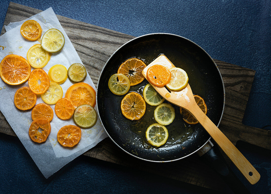Slices of candied orange and lemon in a pan with syrup being lifted out with a wooden spatula to cool on baking paper