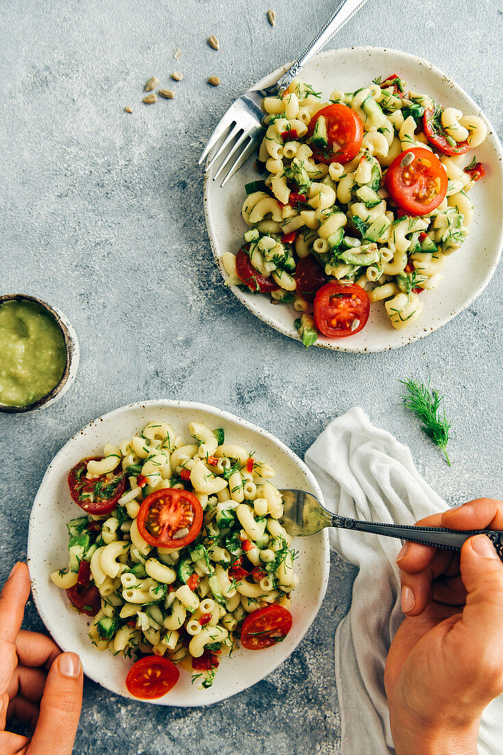 A woman eating vegan pasta salad from a ceramic handmade plate and a small bowl of avocado dressing accompany