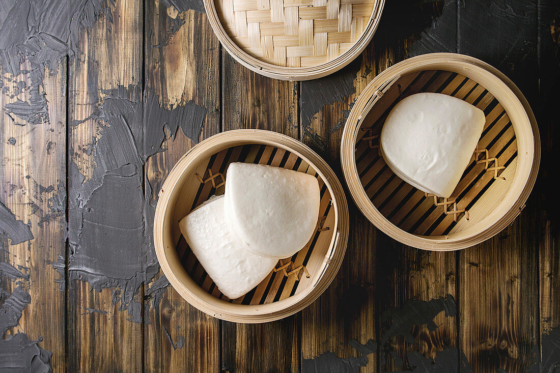 Empty gua bao steamed buns in opened bamboo steamer over dark wooden plank background