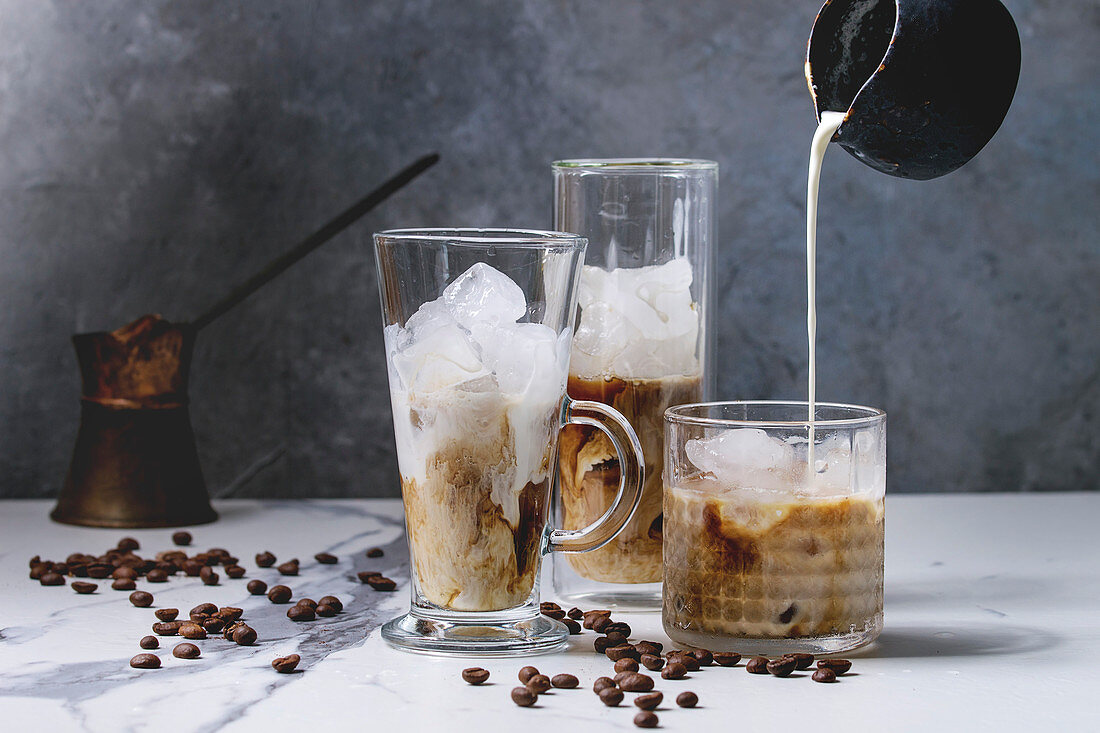 Iced coffee cocktail or frappe with ice cubes and pouring cream in three different glasses with vintage jezva and coffee beans around on white marble table