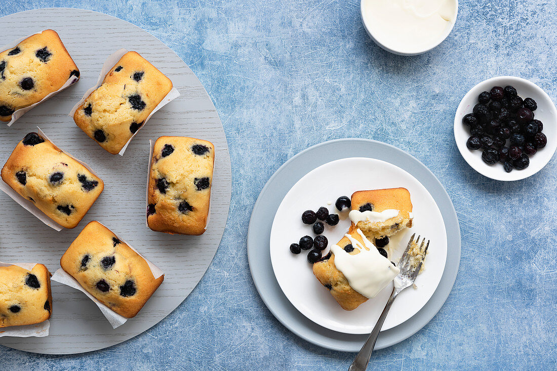 Individual blueberry loaf cakes with a serve on a plate and a bowl of blueberries and a bowl of cream