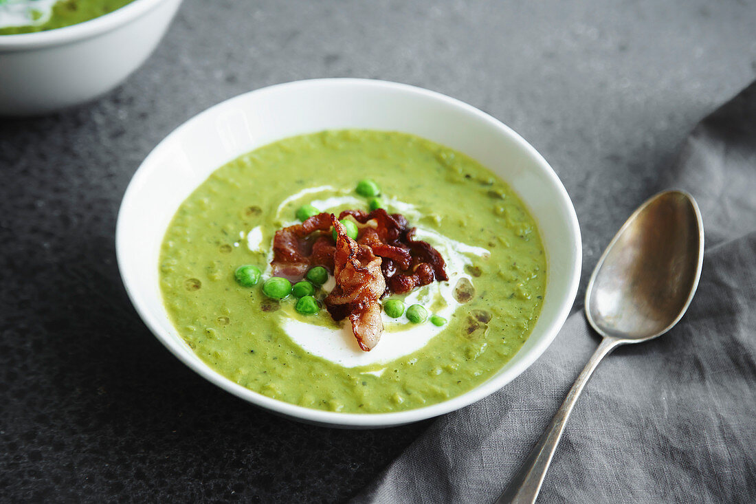 Close up of creamy green pea soup with fried bacon and herbs on gray background