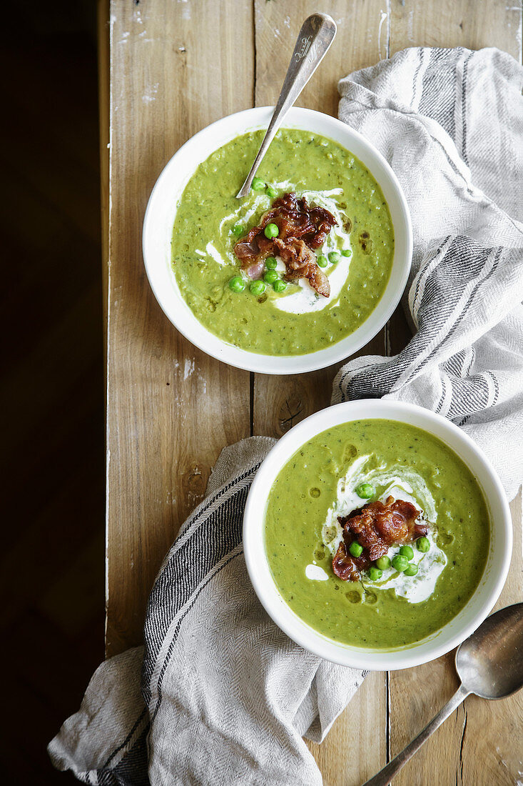 Overhead shot of creamy green pea soup with fried bacon and herbs on rustic wooden background
