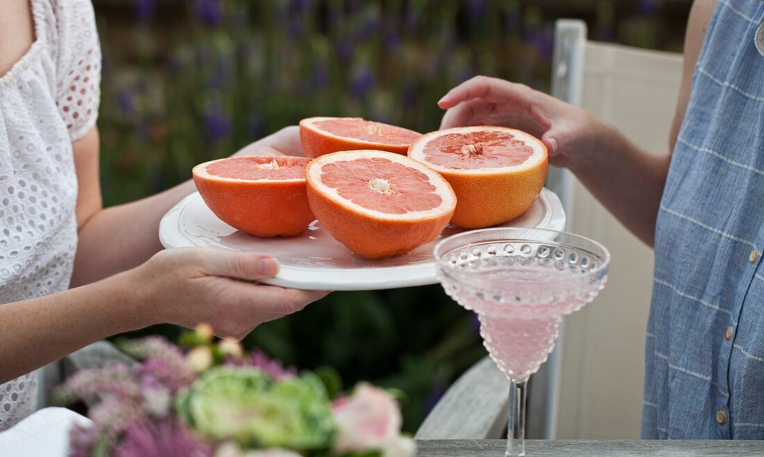 Two women sitting at an outdoor brunch, one woman passing a tray of grapefruit halves and the other woman reaching for one