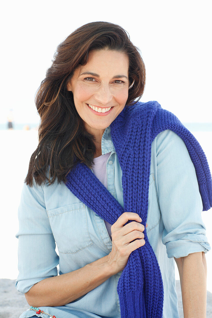 A brunette woman wearing a denim shirt with a knitted jumper over her shoulders