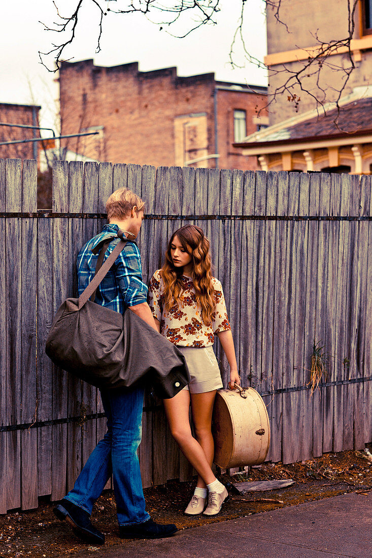 A young couple with luggage standing against a fence