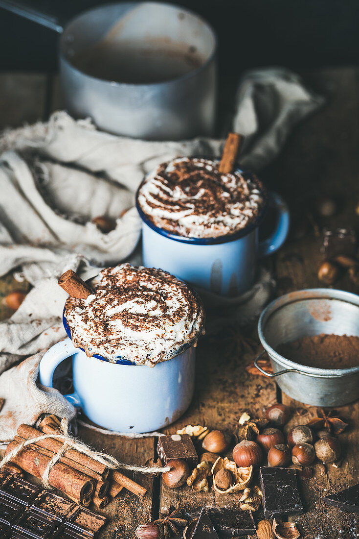 Hot chocolate in blue mugs with whipped cream and cinnamon sticks, spices, nuts and cocoa powder