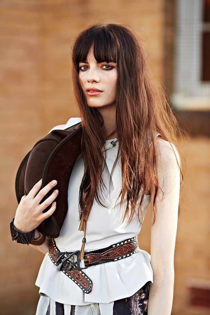 A brunette woman holding a hat and wearing a white top with a belt