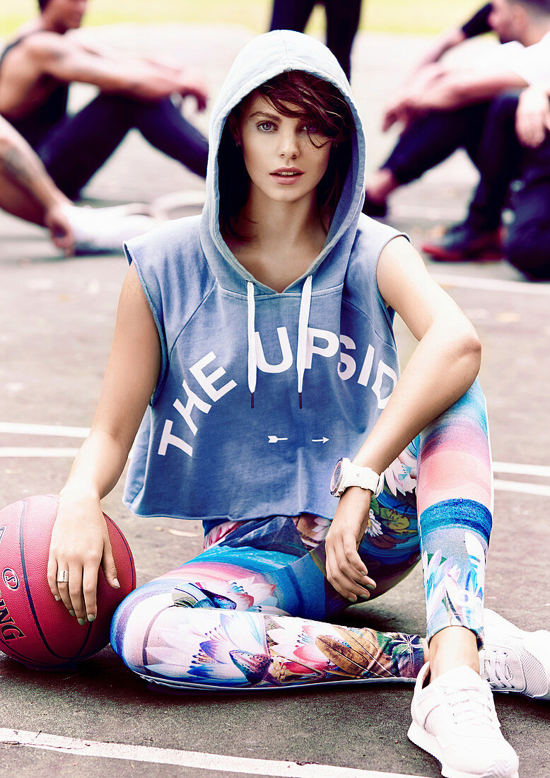Junge Frau in Kapuzenjacke und bunten Leggins mit Basketball auf Betonplatz sitzend