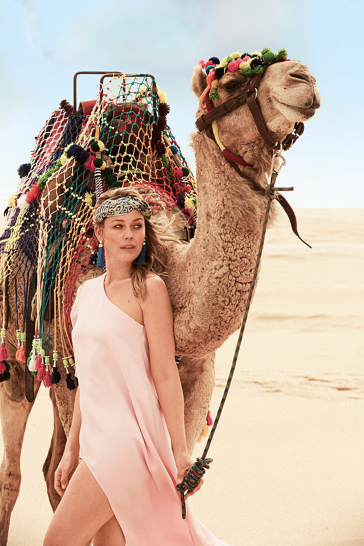 A young woman wearing a pink summer dress with a camel in the desert