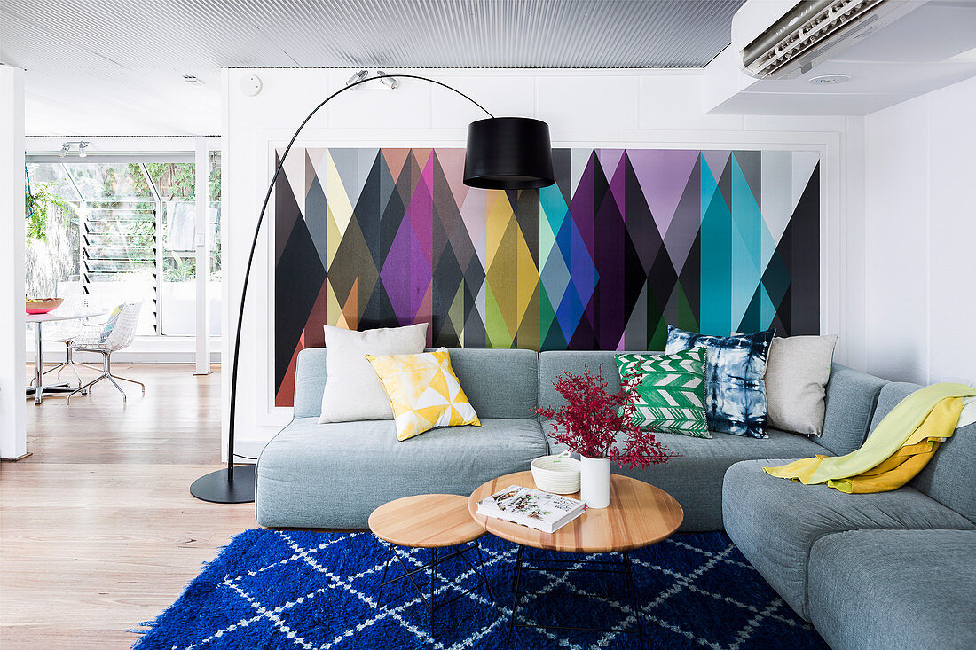 Graues Polstersofa mit Kisssen, Coffeetable und Bogenlampe im Wohnzimmer, an der Wand bunte Tapete mit geometrischem Muster