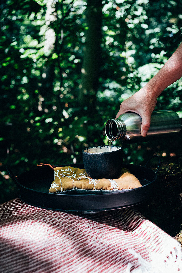 A cup of tea at a tray in the woods, a hand is pouring tea in a cup