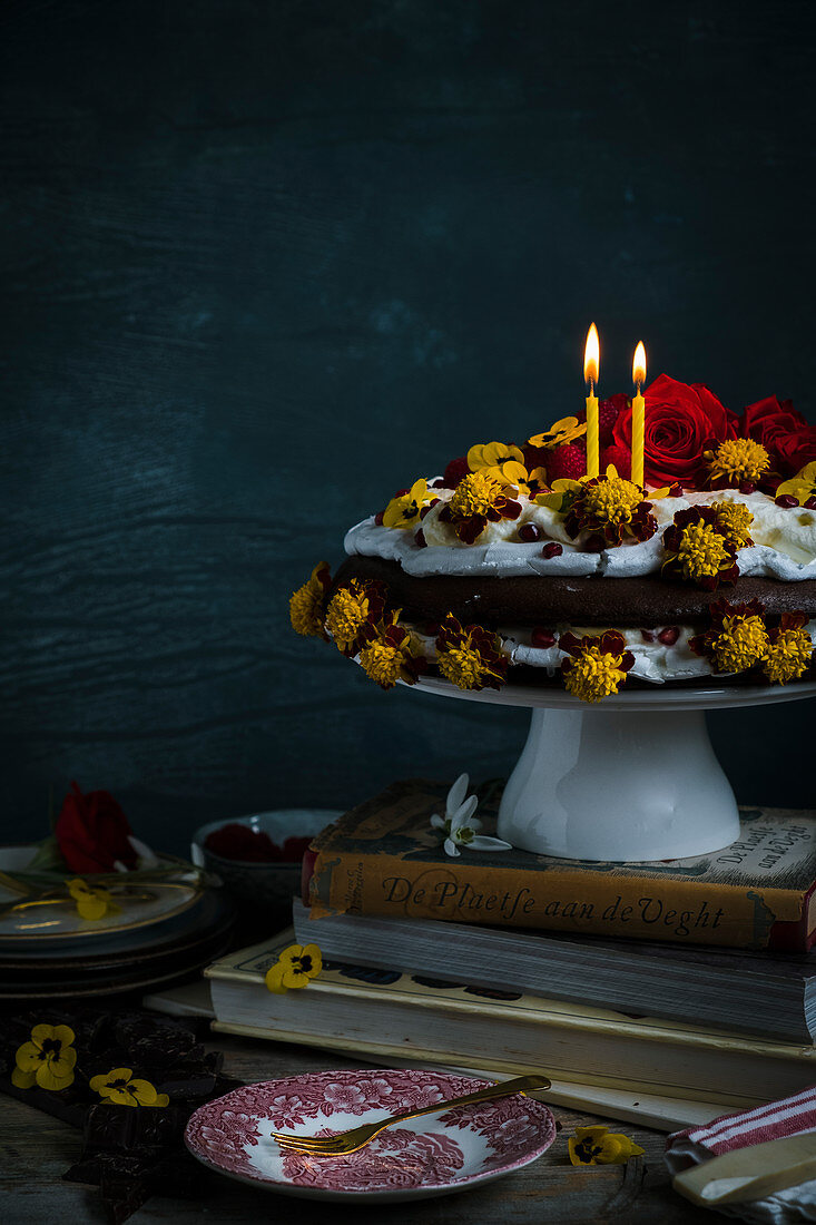 A cake with merengue and chocolate cake with eatable flowers with candles standing on books