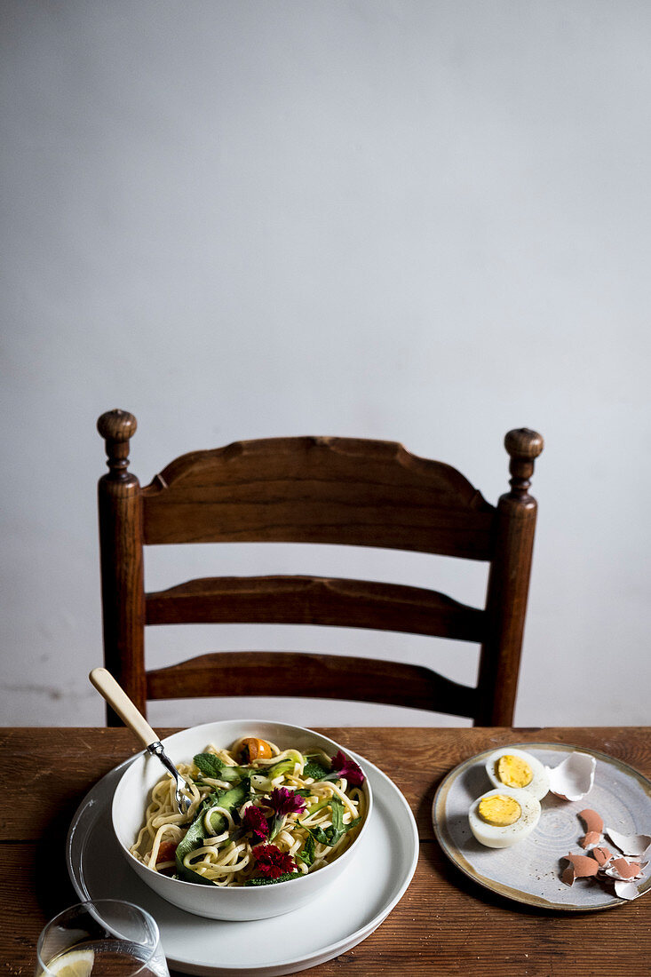 Spaghetti in a bowl on a wooden table with a chair and boiled eggs on a plate