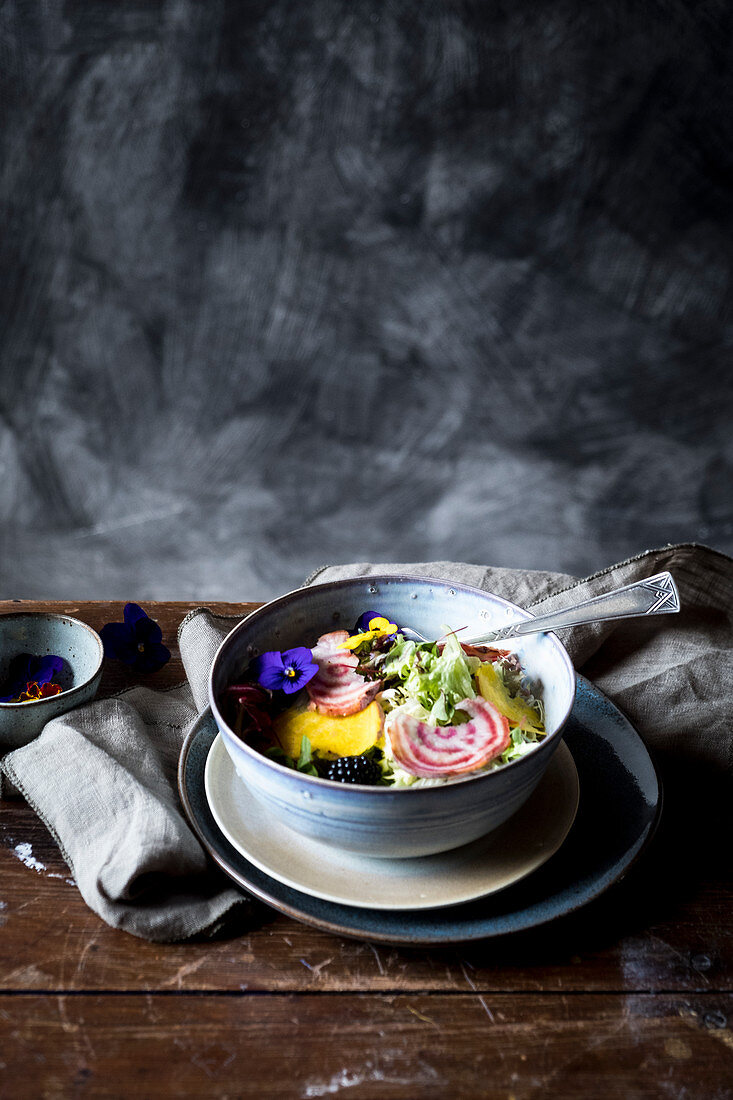 A bowl with salad and eatable flowers at a wooden table from the side