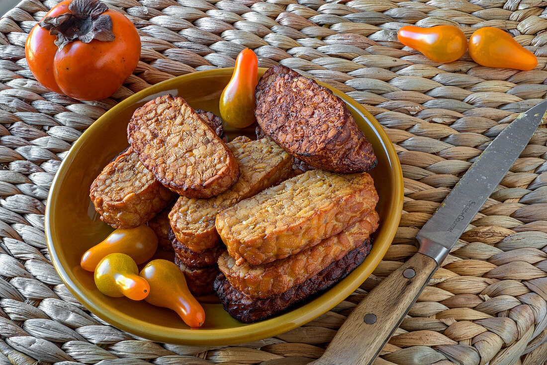 Deep fried tempeh on a braided wicker surface, with bottle tomatoes and a persimmon