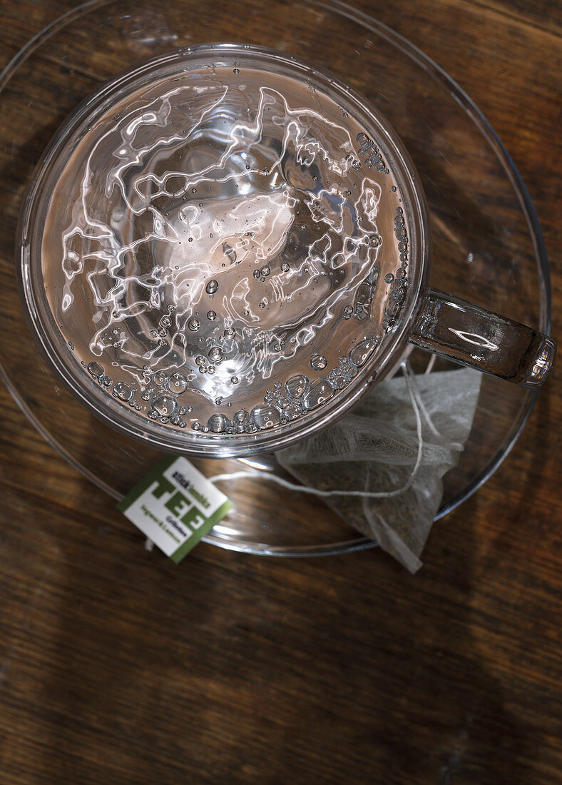 Ein Teeglas mit heißem Wasser, daneben Teebeutel auf Glasteller (Aufsicht)