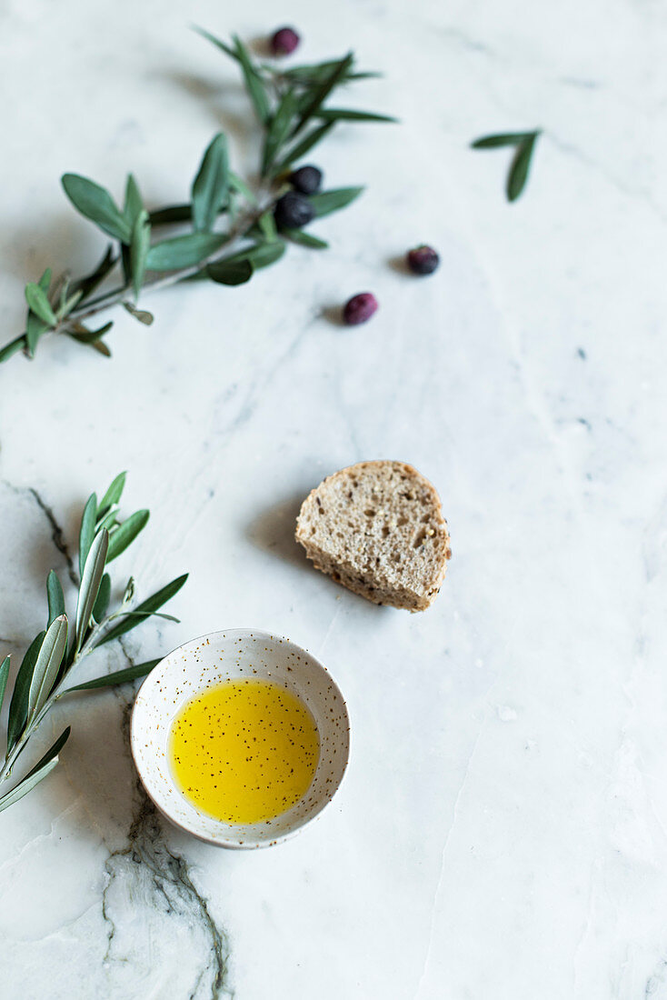 Spanisches Olivenöl in einem Schälchen, Brotscheibe und Olivenzweige auf Marmortisch
