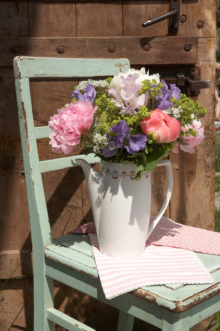 Spring bouquet of peonies, ladies' mantle and sweet peas in coffee pot on wooden chair