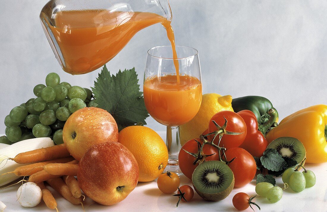 Multivitaminsaft wird ins Glas gegossen, daneben Obst, Gemüse