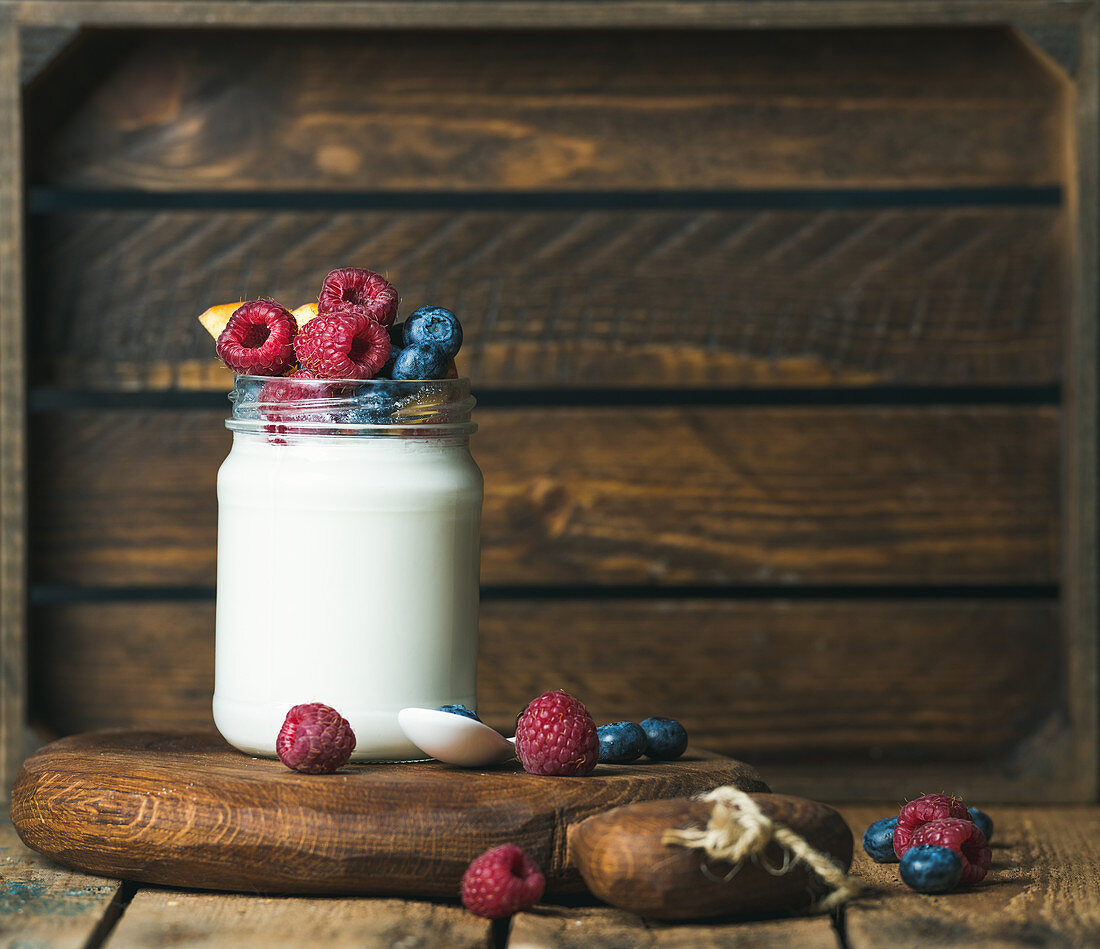 Glass jar of white yogurt with fresh garden berries and peach on serving board over rustic table