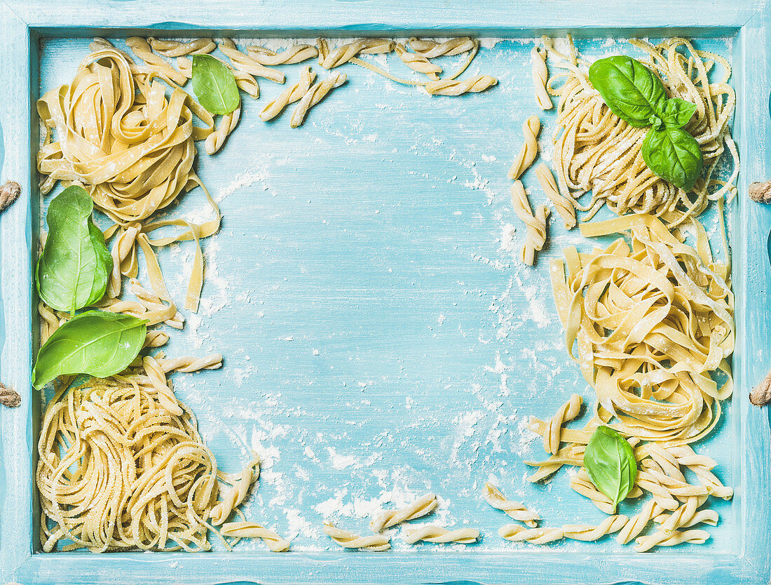Various homemade fresh uncooked Italian pasta with flour and green basil leaves in blue wooden tray
