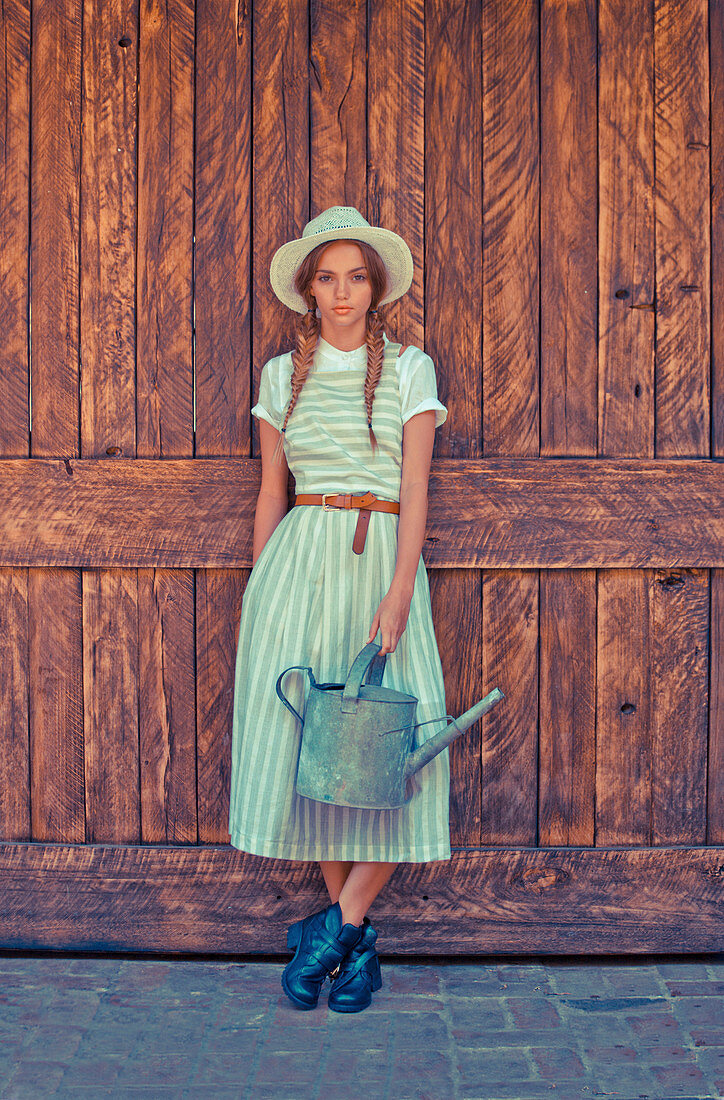 A young woman with a watering can wearing a blouse and a striped pinafore dress