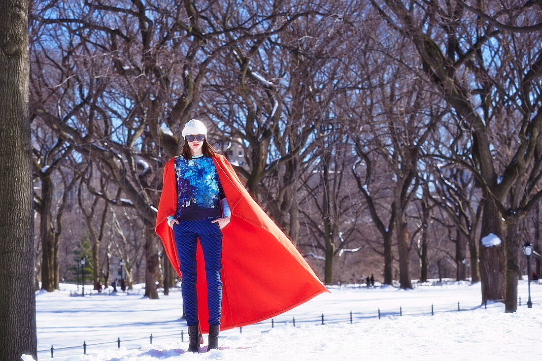 Brünette Frau in rotem Cape, blauem Shirt und blauer Hose in winterlichem Park