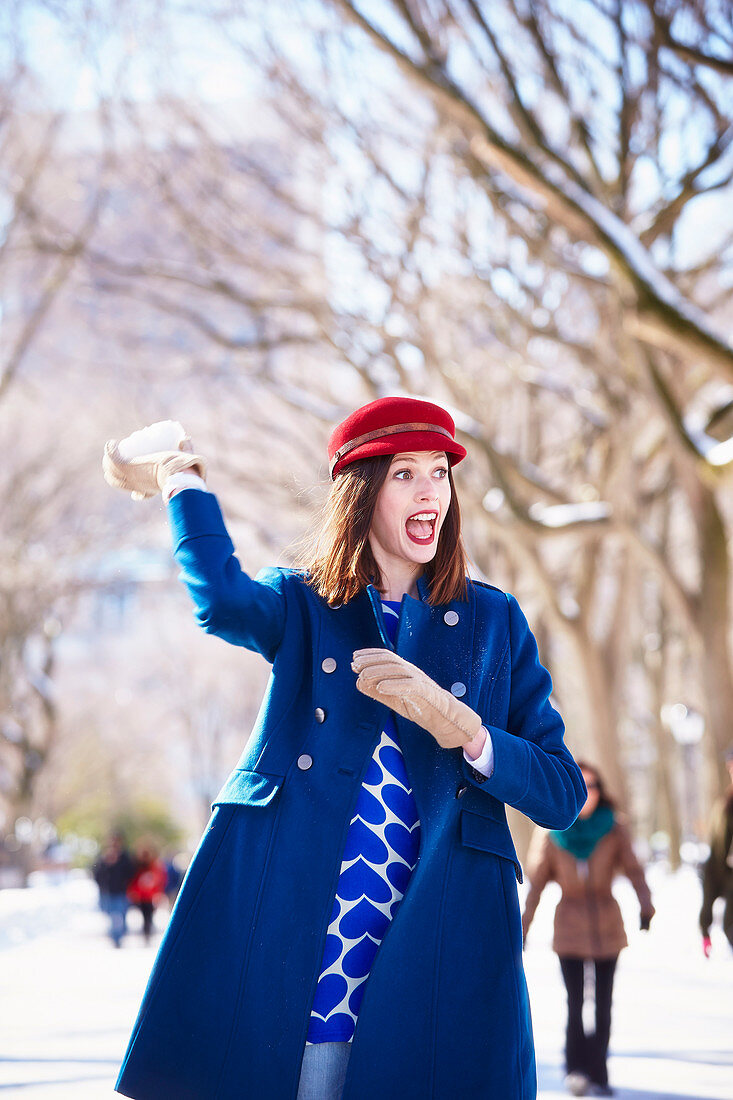 Brünette Frau mit rotem Hut und Schneeball in blauem Mantel in winterlichem Park