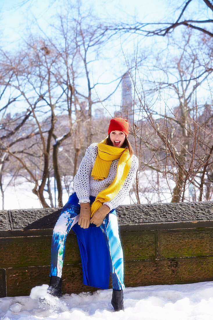 Junge Frau mit roter Mütze und gelbem Schal in weißem Strickpulli und blauer Hose in verschneitem Park