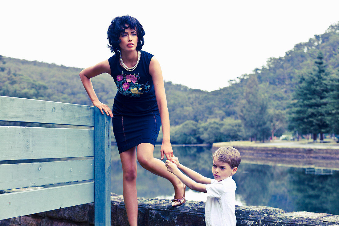 A dark-haired woman wearing a dark blue dress with floral appliqué with a boy by a lake
