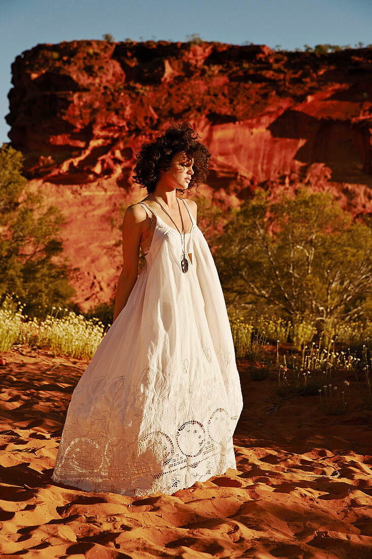 A dark-haired woman wearing a long, white, embroidered summer dress in a desert