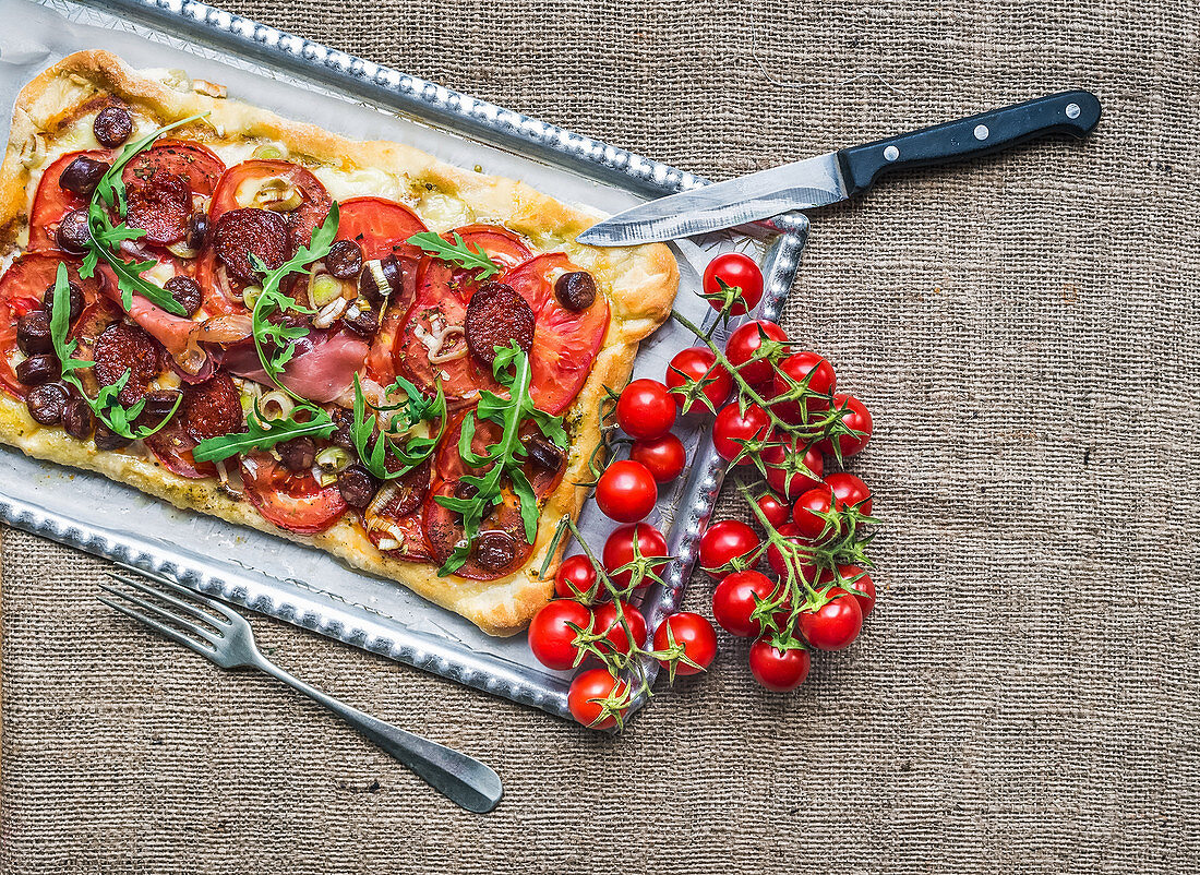 Homemade square pizza with meat, salami, cherry-tomatoes and fresh arugula on a silver tray