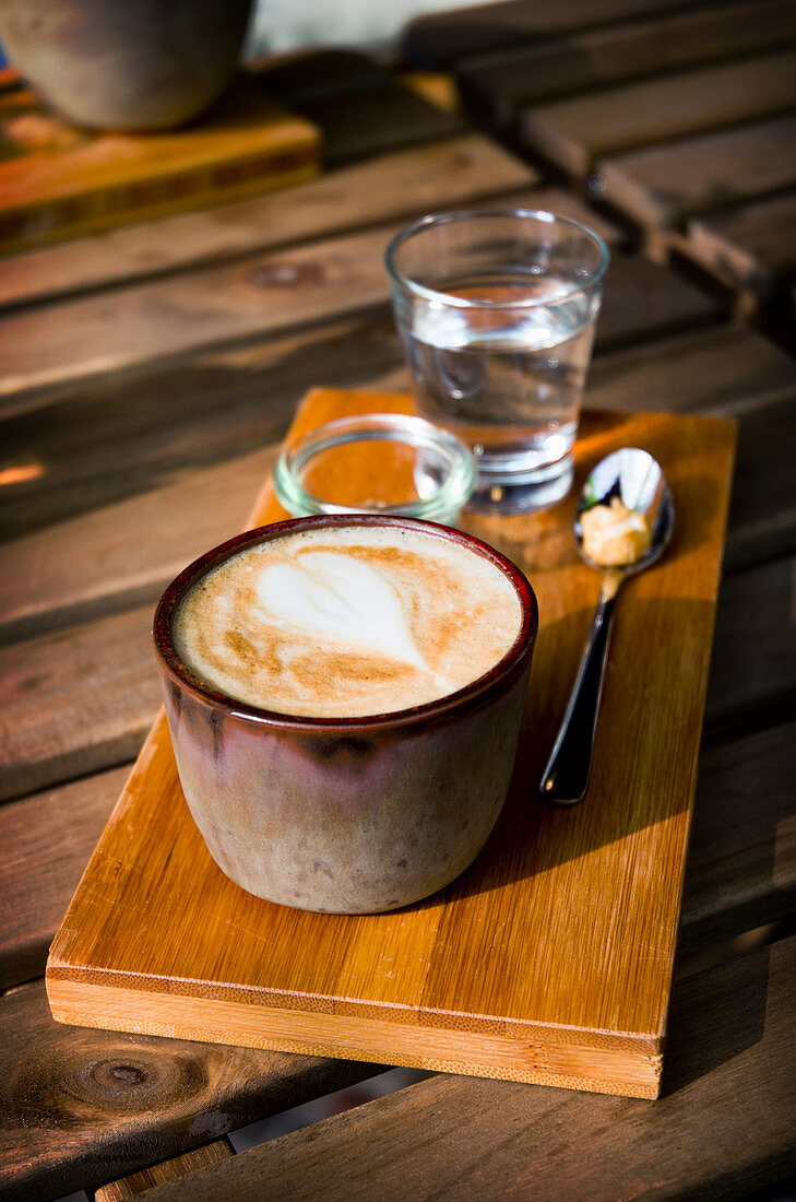 A cappucino in a rustic earthenware cup on a wooden board with a glass of water and sugar