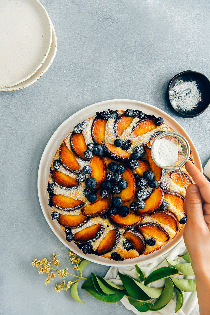A woman sifting powdered sugar on a peach cake with blueberries