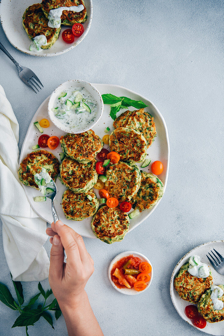 Hand spreading cucumber yogurt dip on zucchini fritters served on a white ceramic plate