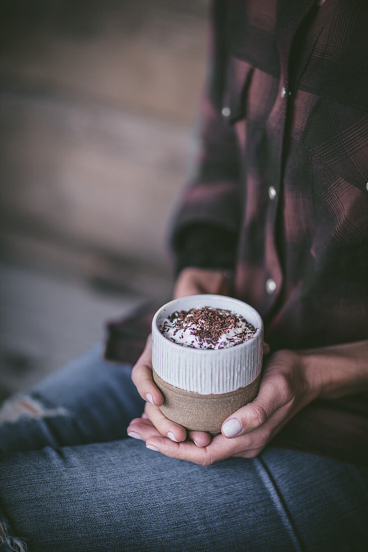 A hand held cup of hot chocolate with whipped cream and chocolate sprinkles, served in a white and tan ceramic mug
