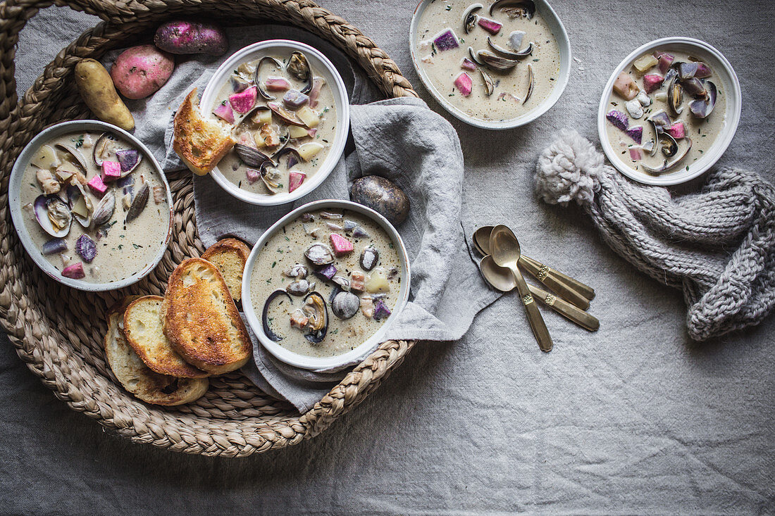 Five bowls of clam chowder with colorful heirloom potatoes served in white dishes and a straw tray