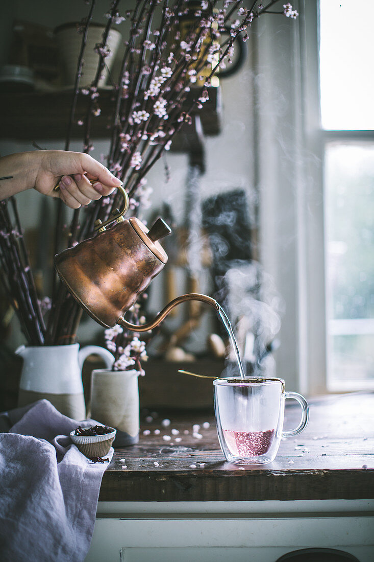 Hibiscus tea being poured from a copper kettle into a glass mug on a rustic wood kitchen countertop