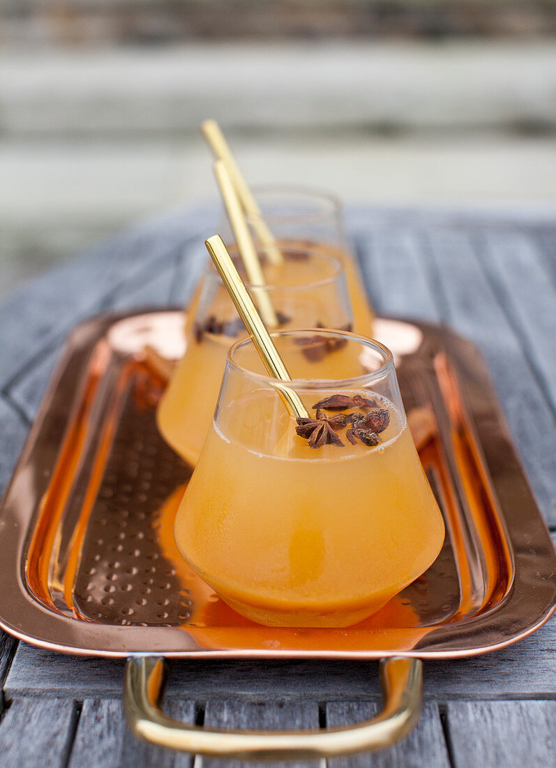 A copper tray with apple cider, with star anise and gold straws, sitting on an outdoor table with stone steps in the background