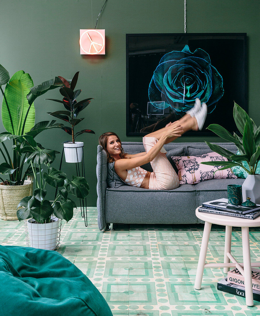 Young woman lying on green upholstered sofa in living room with house plants and green wall