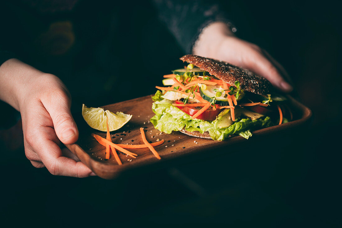 Woman holding a tray with a vegetarian sandwich with lettuce, cucumber, carrot tomato anda avocado