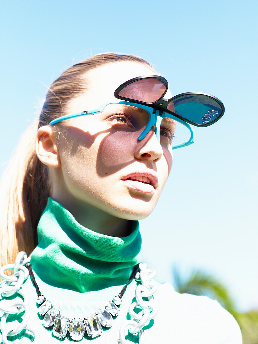 Junge blonde Frau mit hochklappbarer Sonnenbrille und grünem Turtleneck Pullover schaut in die strahlende Sonne