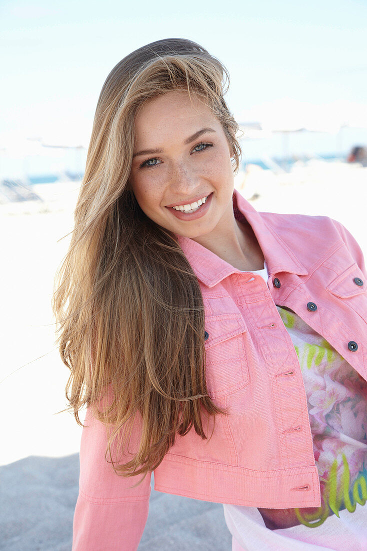 A young blonde woman on a beach wearing a printed t-shirt and a pink denim jacket