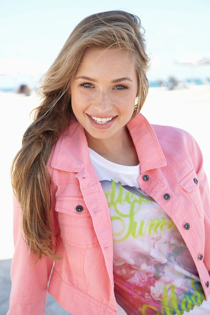 Junge blonde Frau in bedrucktem T-Shirt und rosa Jeansjacke am Strand